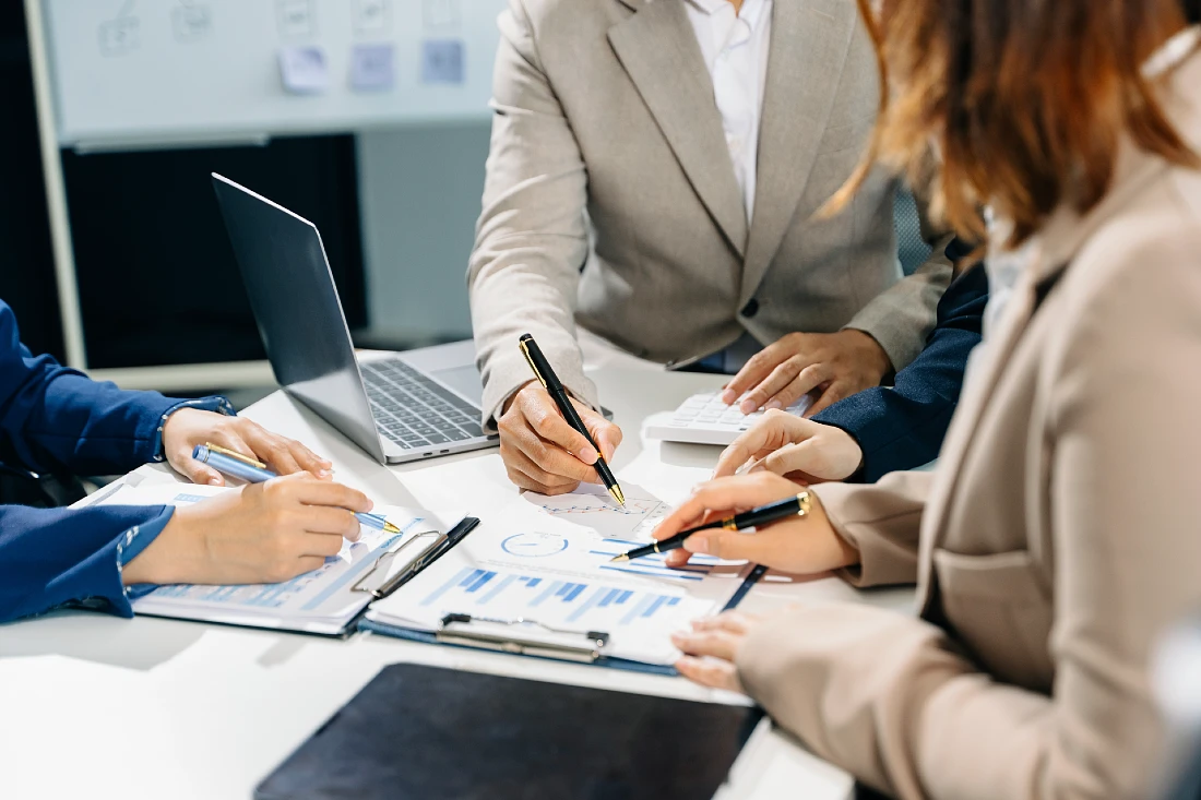 three people are working together at a table, all of them holding pens and pointing at a document